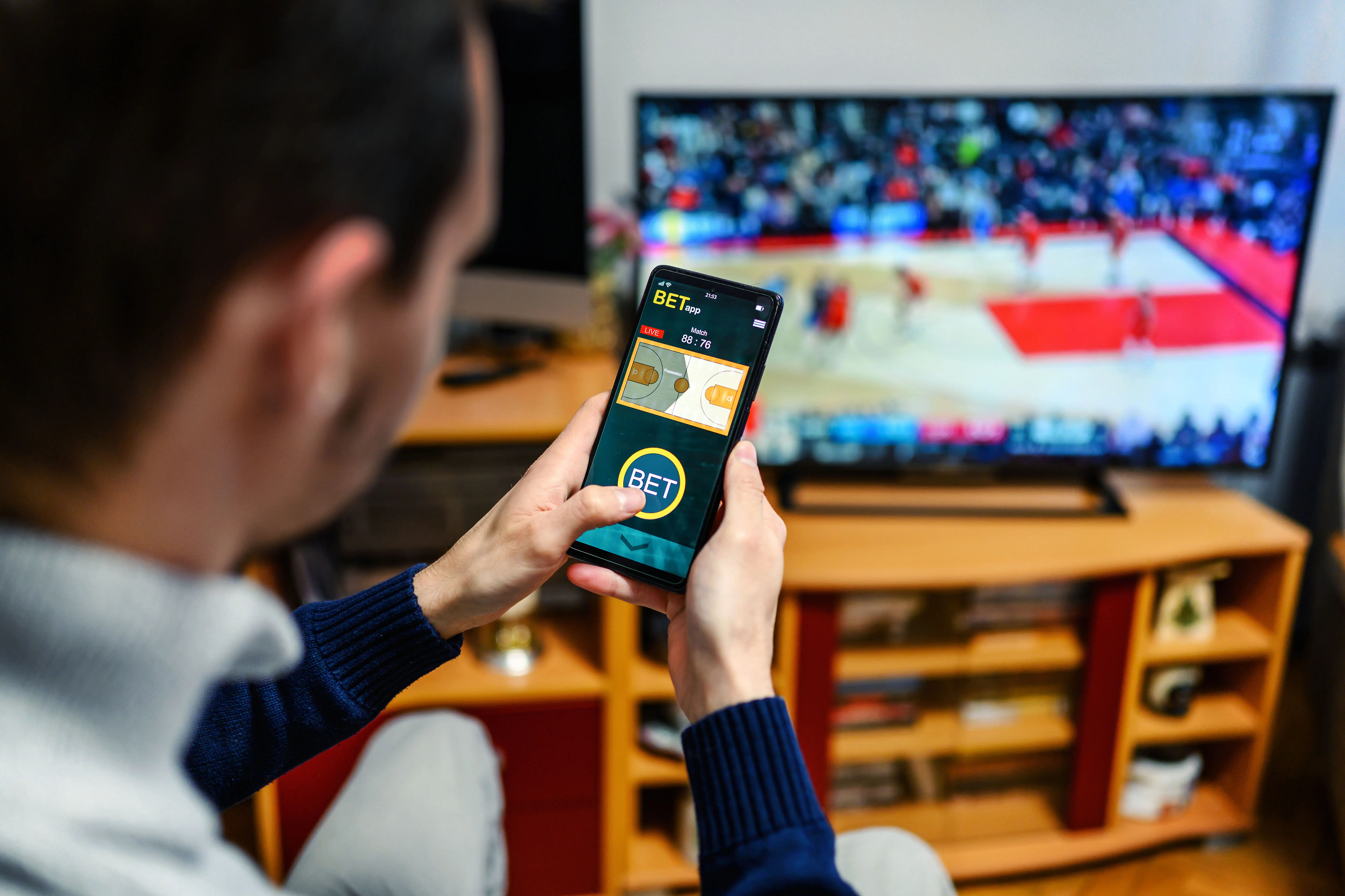 Young men are sitting on the sofa at home and watching a basketball game. Use your smartphone for sports betting.
