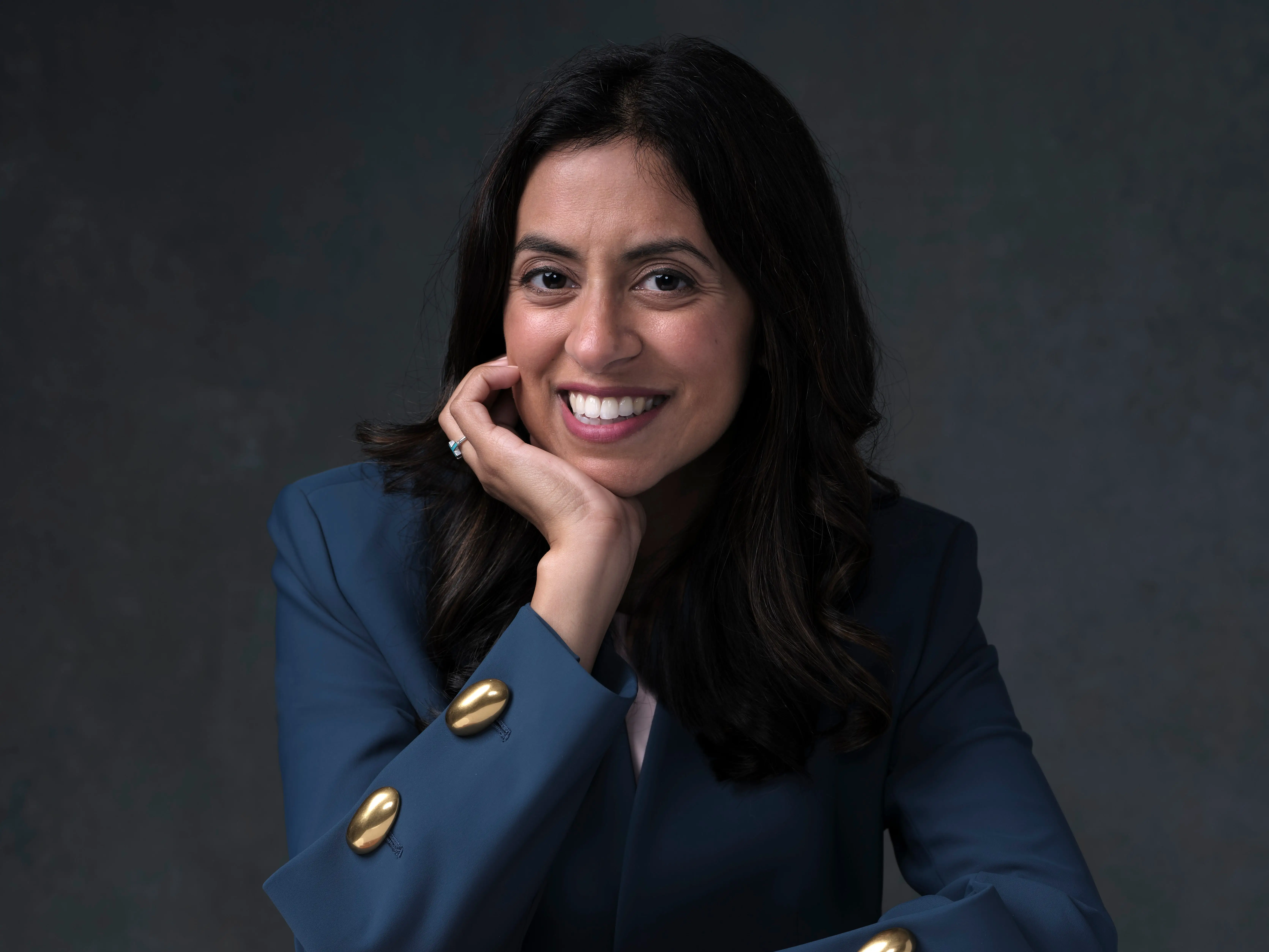 Dr. Trisha Pasricha in a dark blue blazer is sitting at a table and smiling with her chin propped on her hand