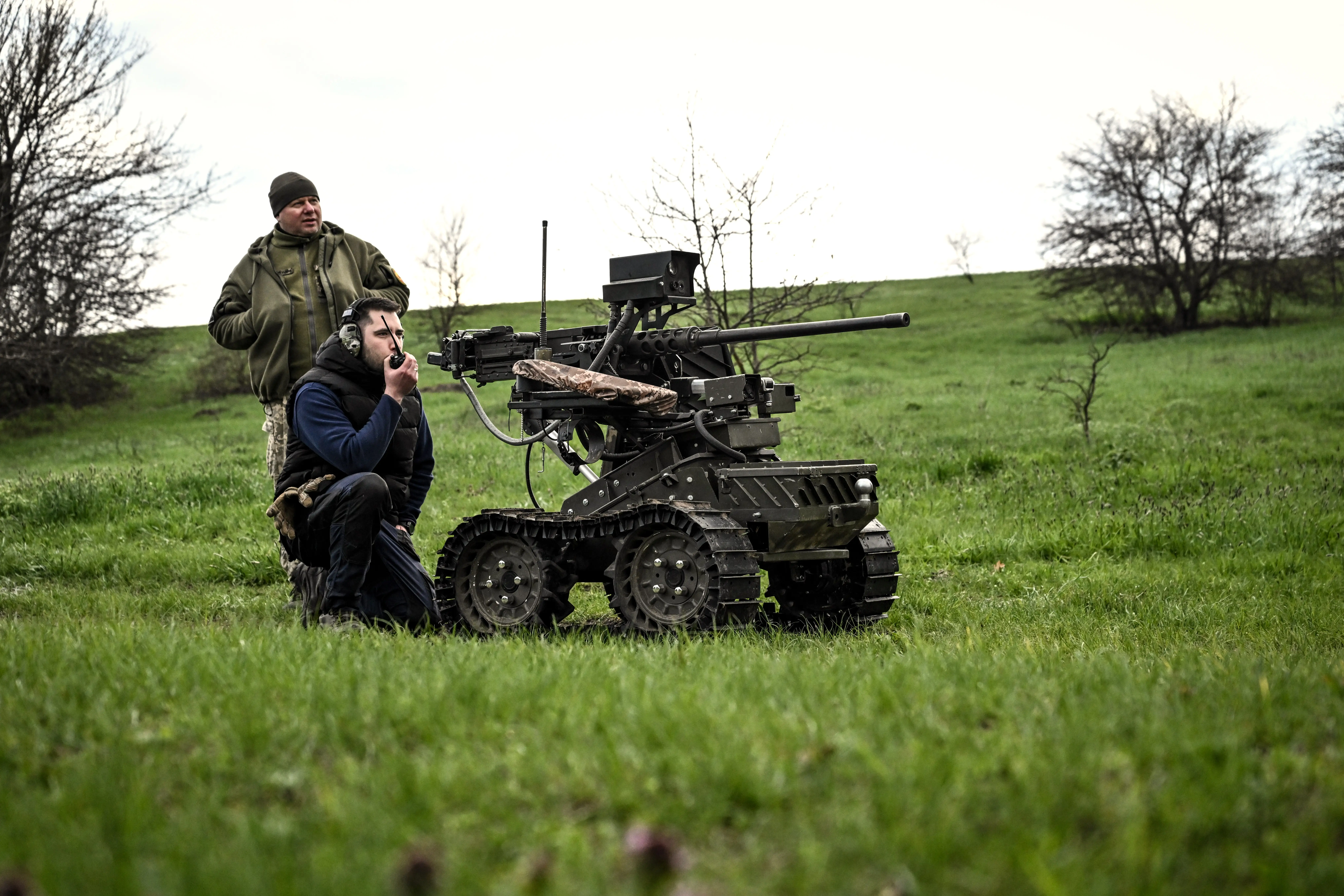 A robot on tracks in a grassy field with two men beside it