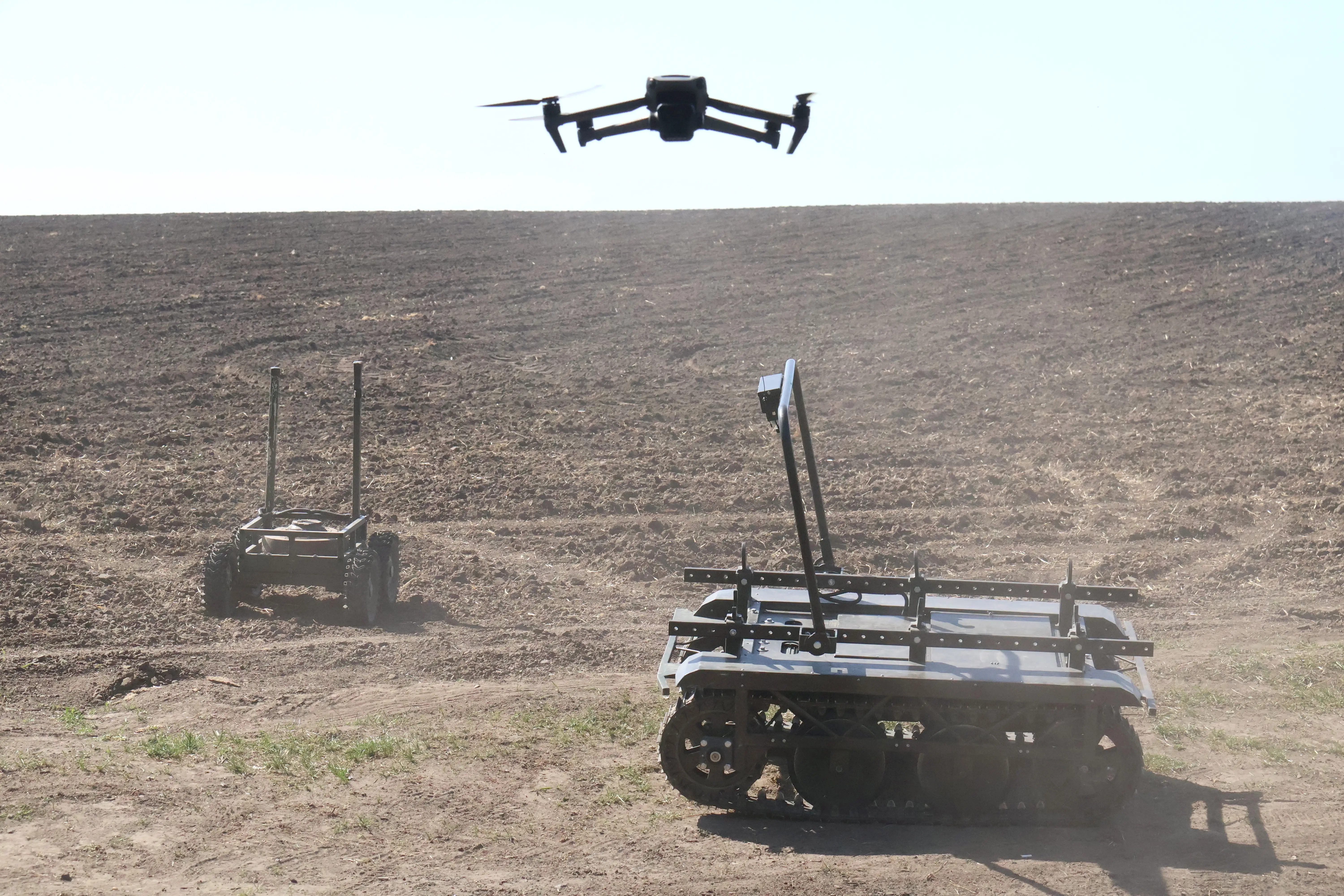 Two ground robots on dusty brown ground with a drone flying above them
