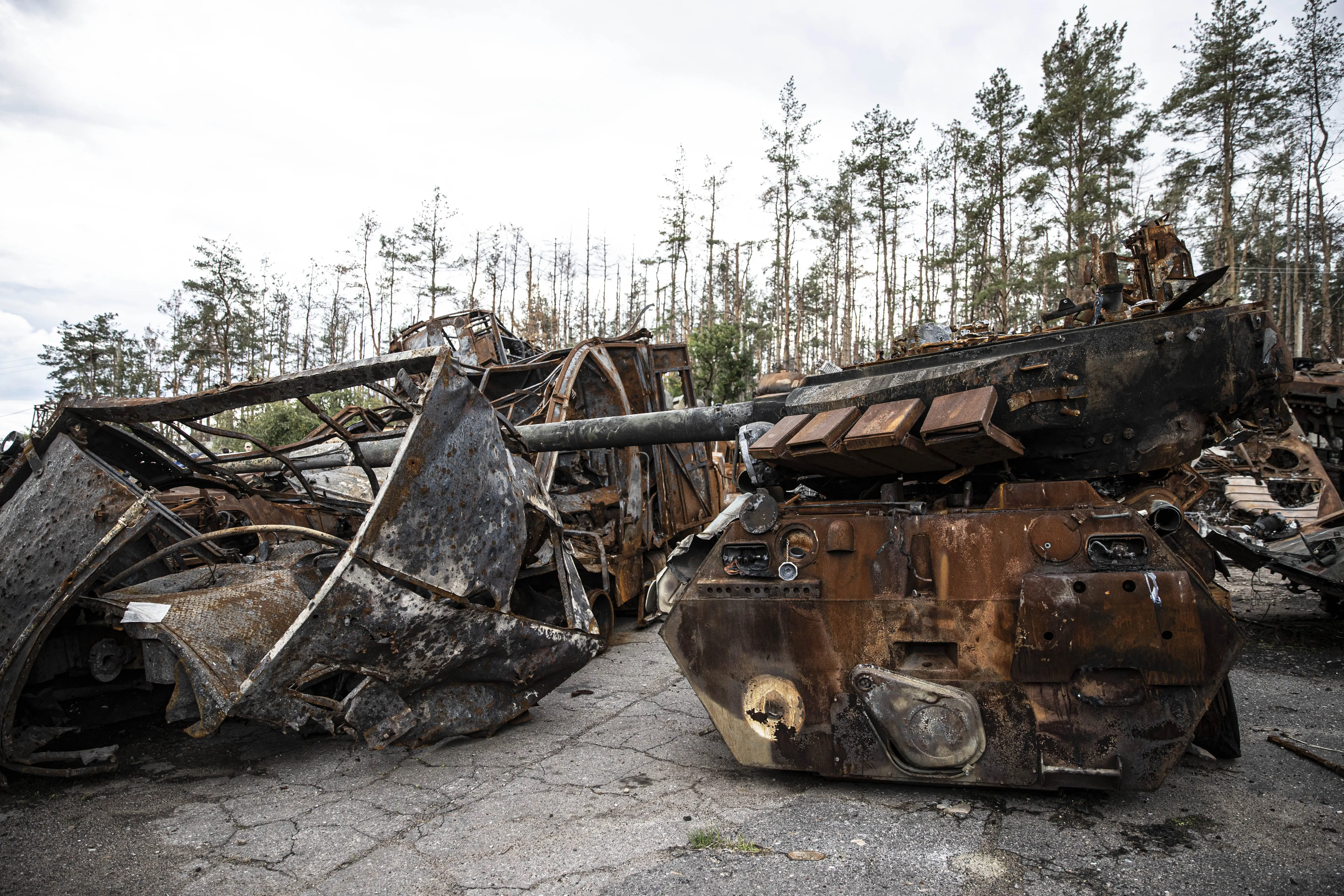 Damaged and rusted tank parts on tarmc under a grey sky and in front of some trees