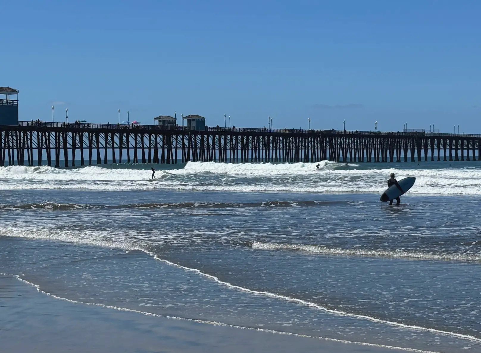 A surfer holding a surfboard at the edge of the Oceanside beach, with the boardwalk in the background.