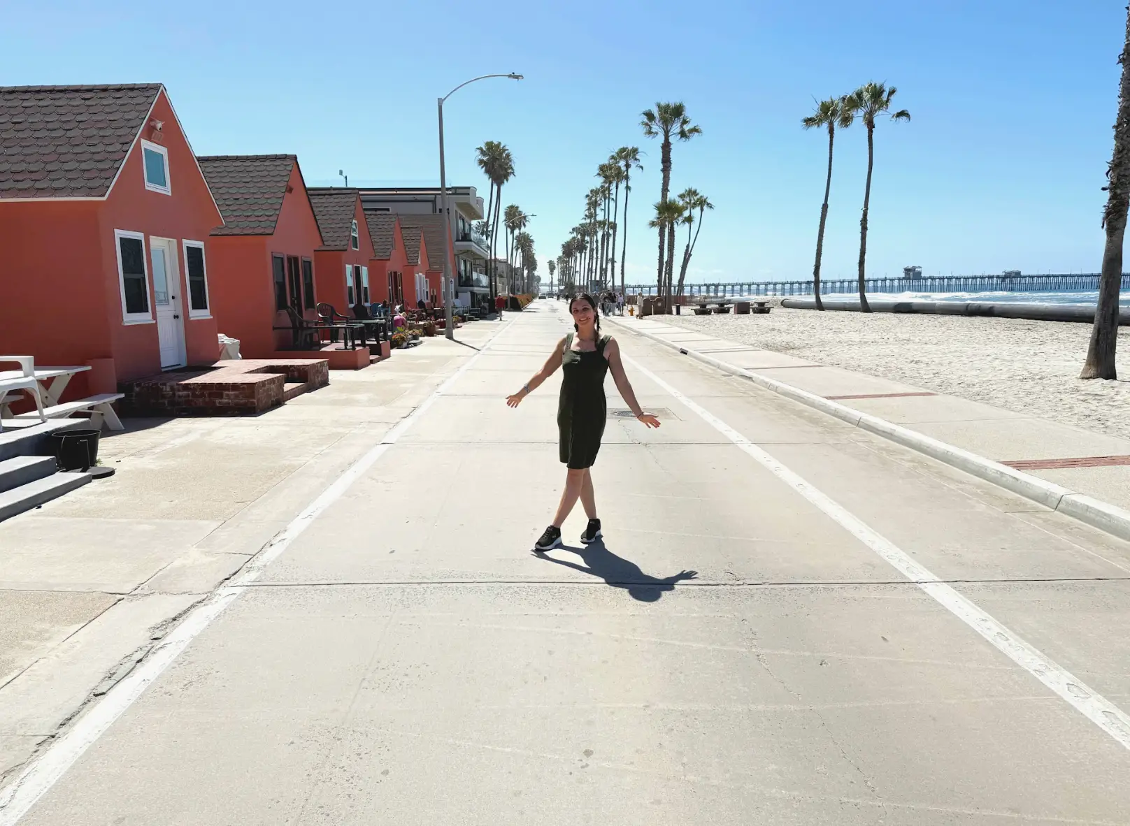 The writer posing in the middle of the street next to a beach in Oceanside, California, with trees and small houses in the background.