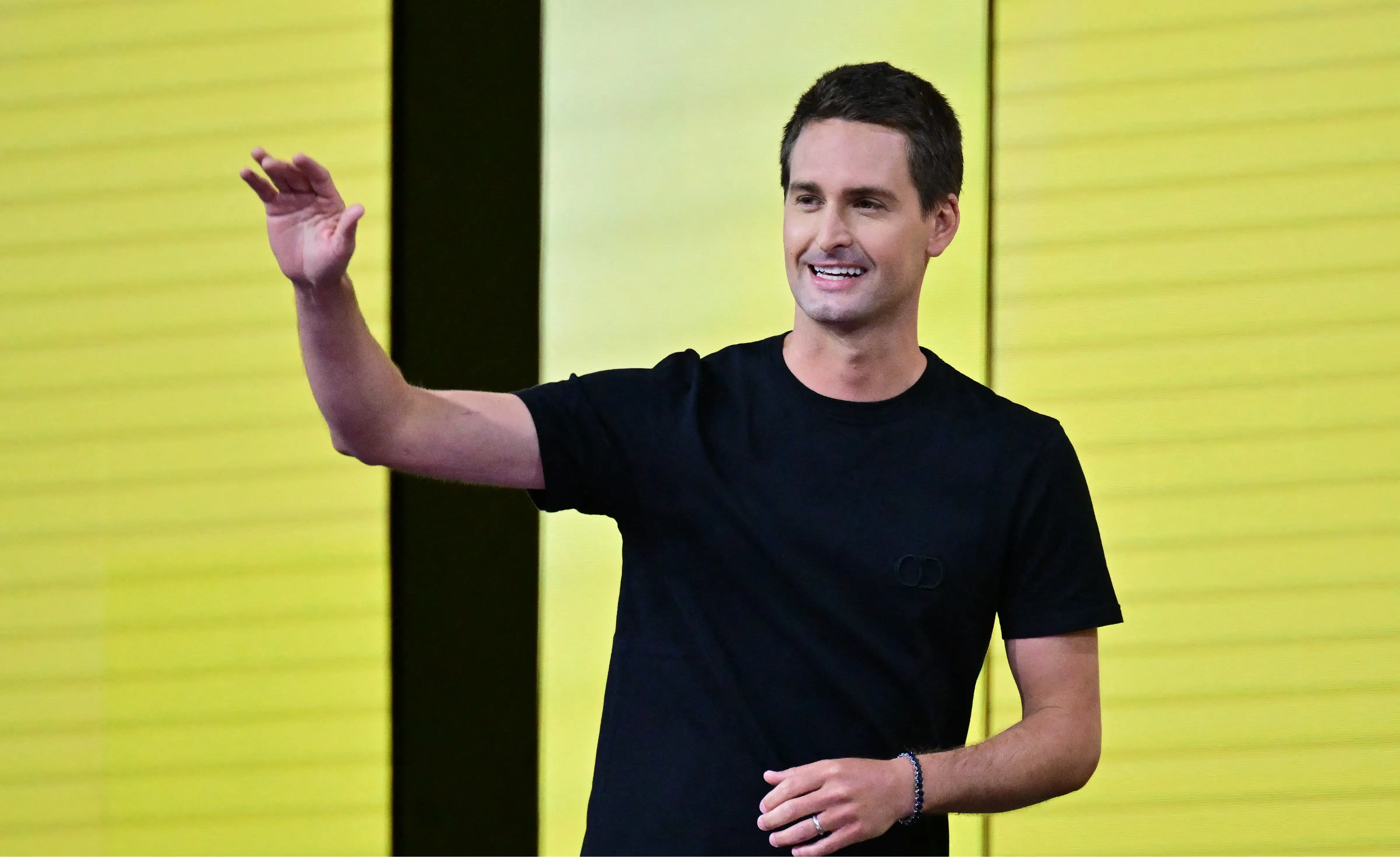 Evan Spiegel, wearing a black shirt and a wristband, waves while standing in front of a bright yellow wall.