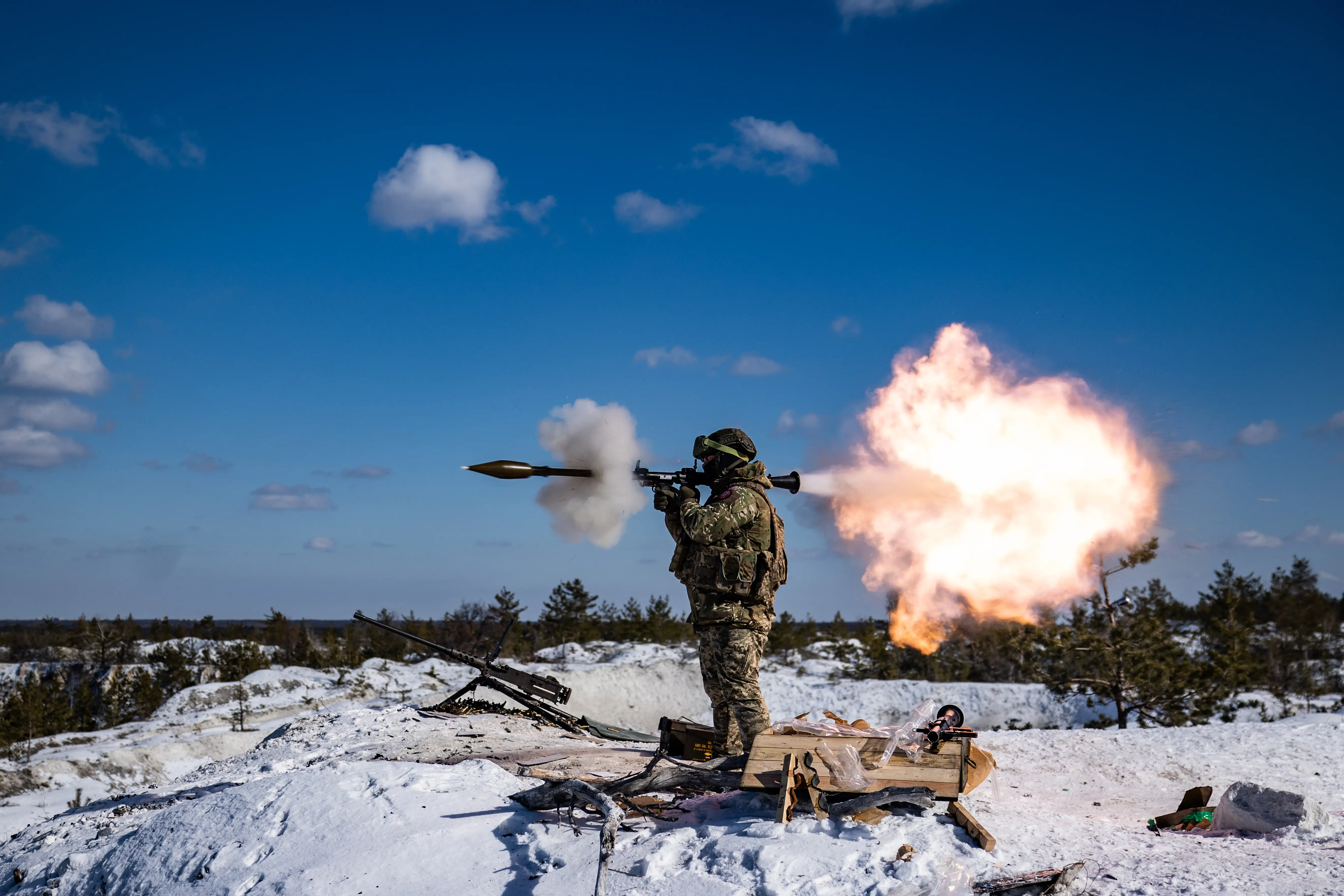 A man in camouflage gear fires a shoulder weapon in snow under a blue sky