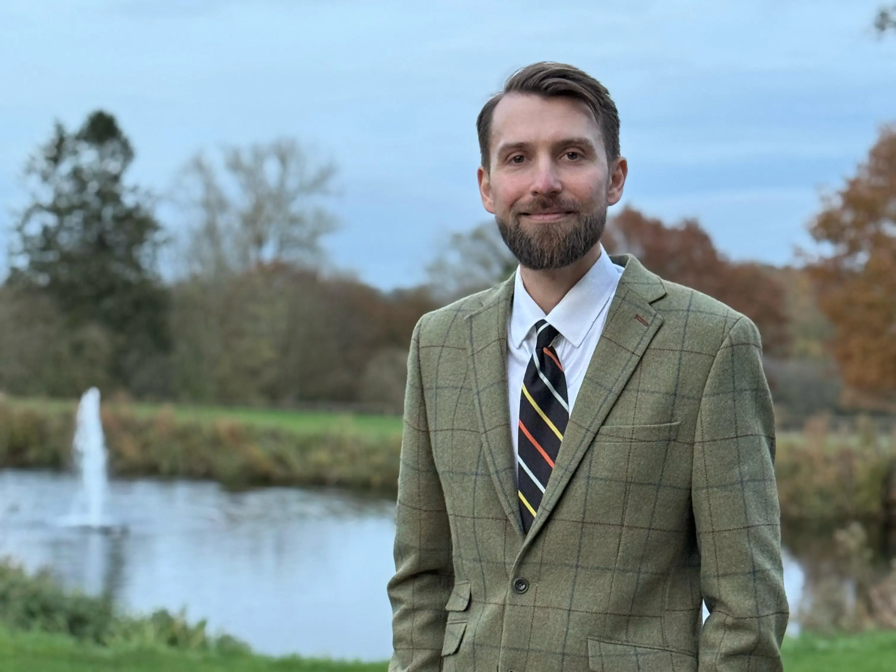 A young man in a tweed-style blazer smiles by a lake.