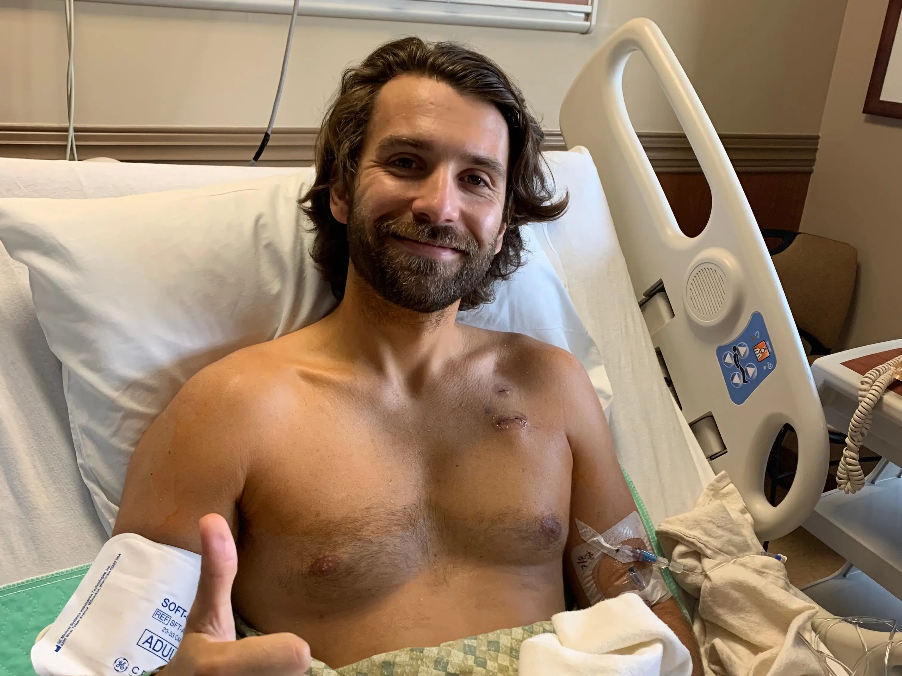 A young man lying in a hospital bed gives a thumbs-up sign.