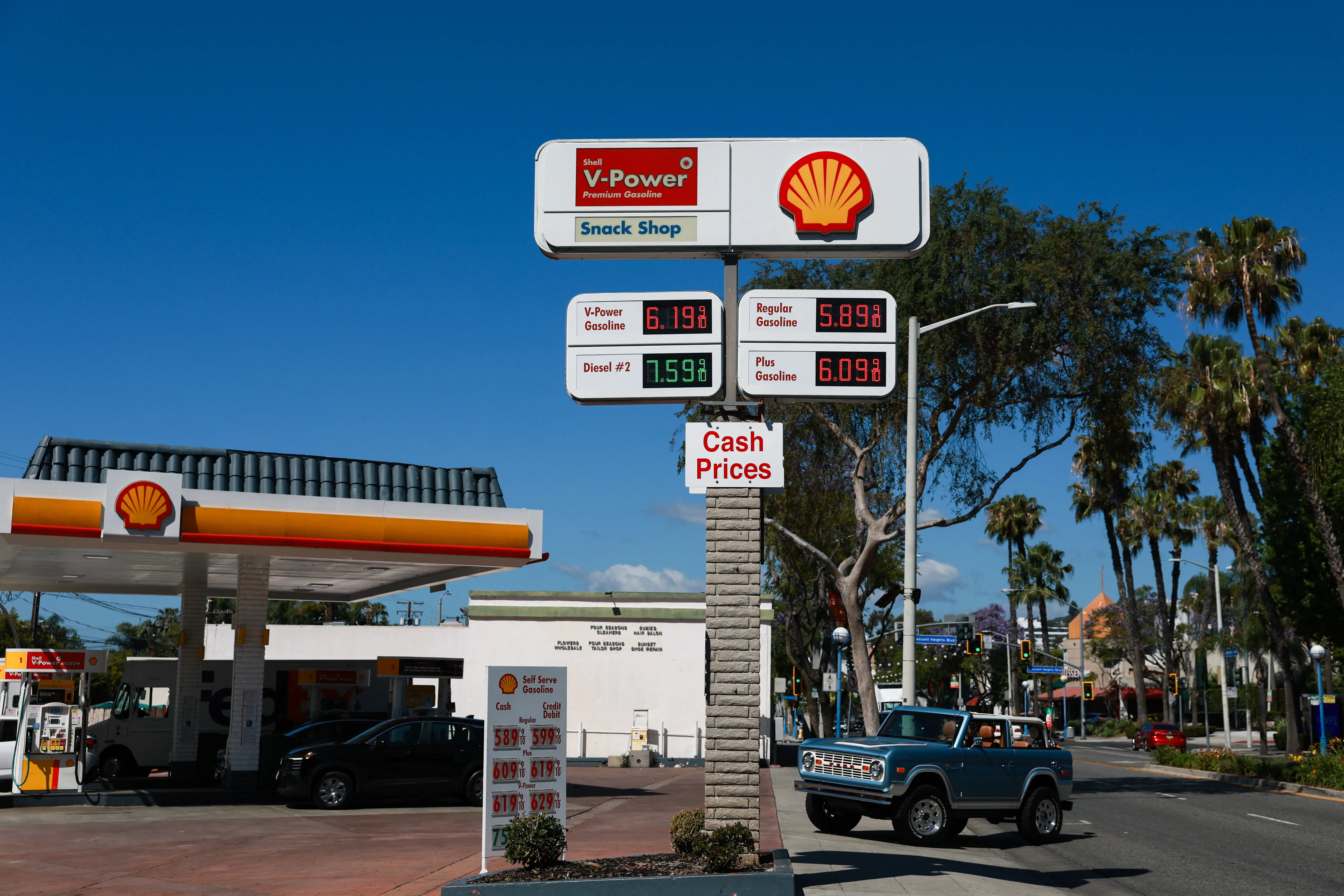 A blue Ford Bronco drives past palm trees into a gas station lot. The price for a gallon of regular gas at the station is $5.89.