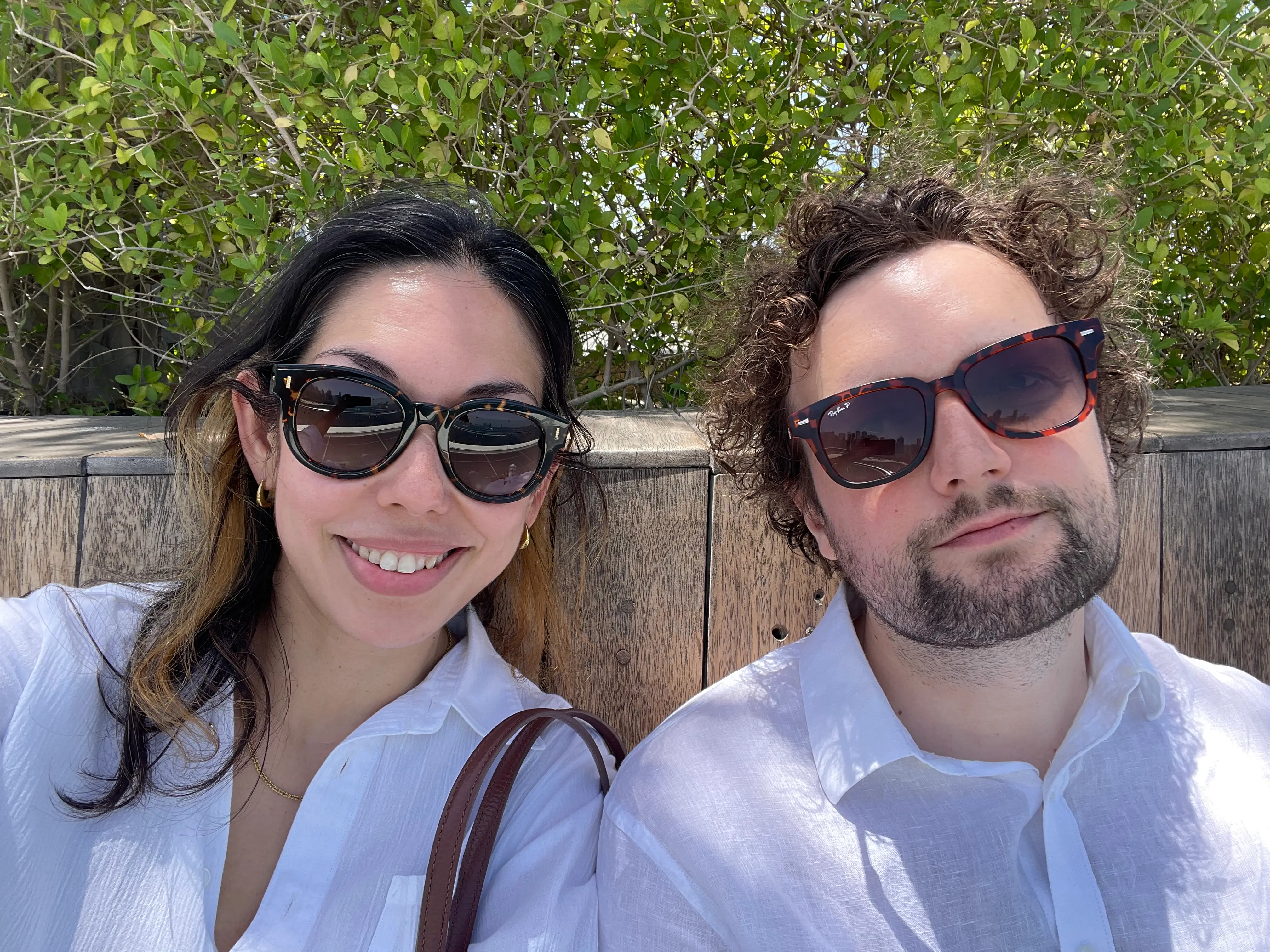 A couple wearing sunglasses and bushes behind them.