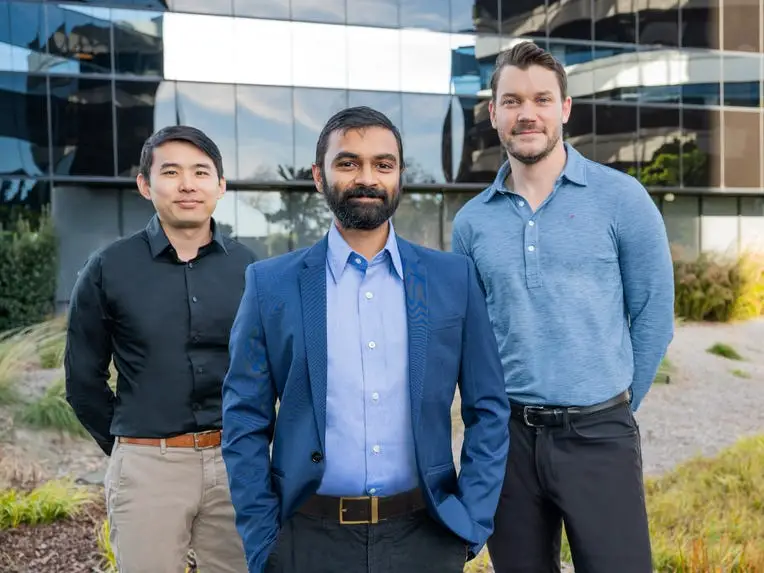 Three men pose outside a glass office building.