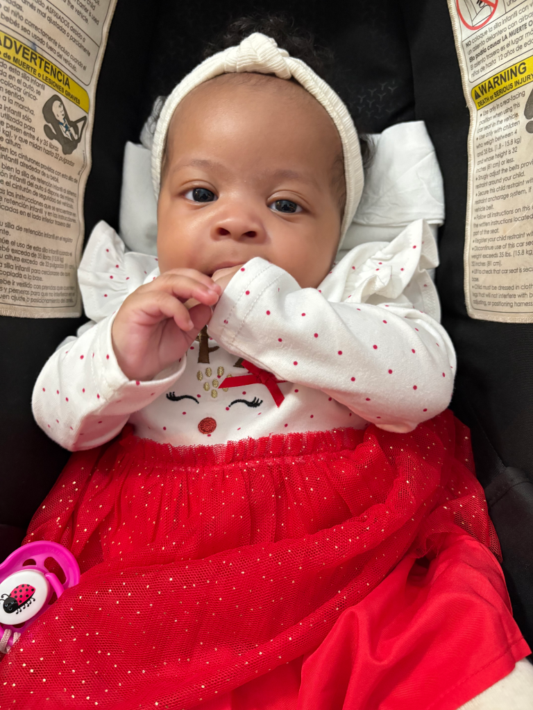 A baby girl in a car seat, wearing a white shirt with red polka dots and a red skirt, with a matching white headband.