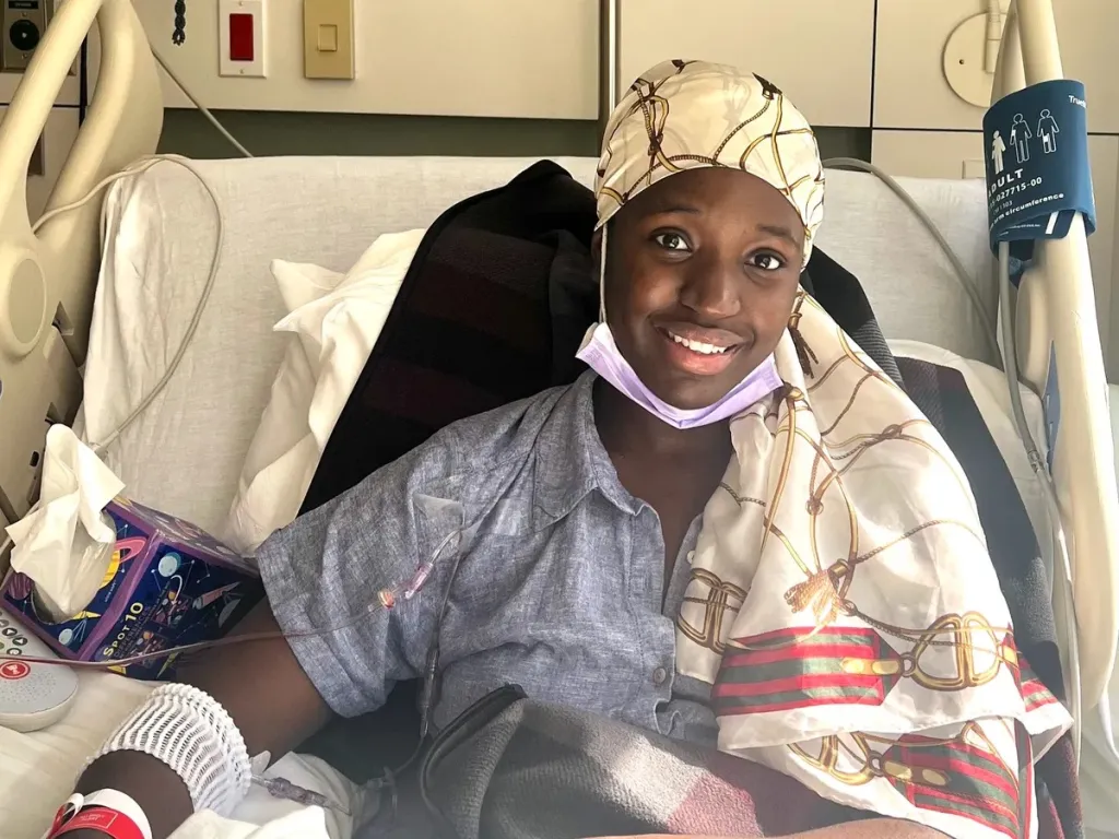 A smiling young woman in a headscarf sits in a hospital bed with an IV in her arm.