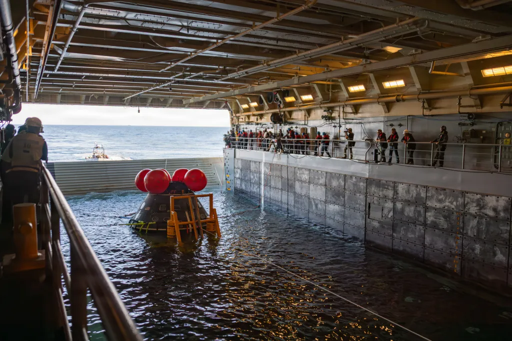 An Orion test capsule floats in the ocean within the well deck of the USS John P. Murtha as crew members observe from above.