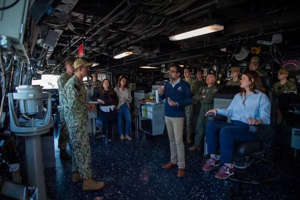 A group of people, including sailors and civilians, gather in a ship's interior as one man speaks.