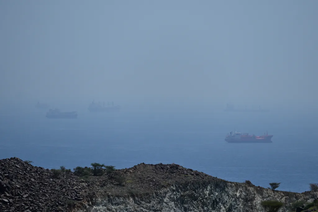 A hazy view of several cargo ships in the distance on a blue ocean, seen from a rocky hillside.
