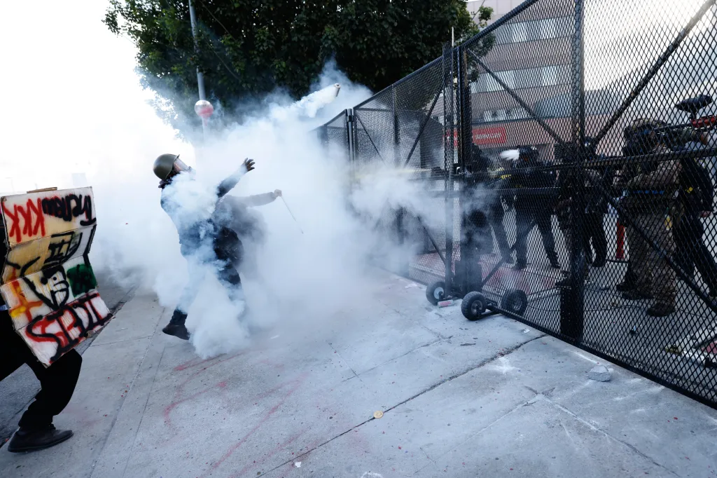 A protester in a helmet throws a smoke grenade as law enforcement officers behind a fence spray a deterrent.