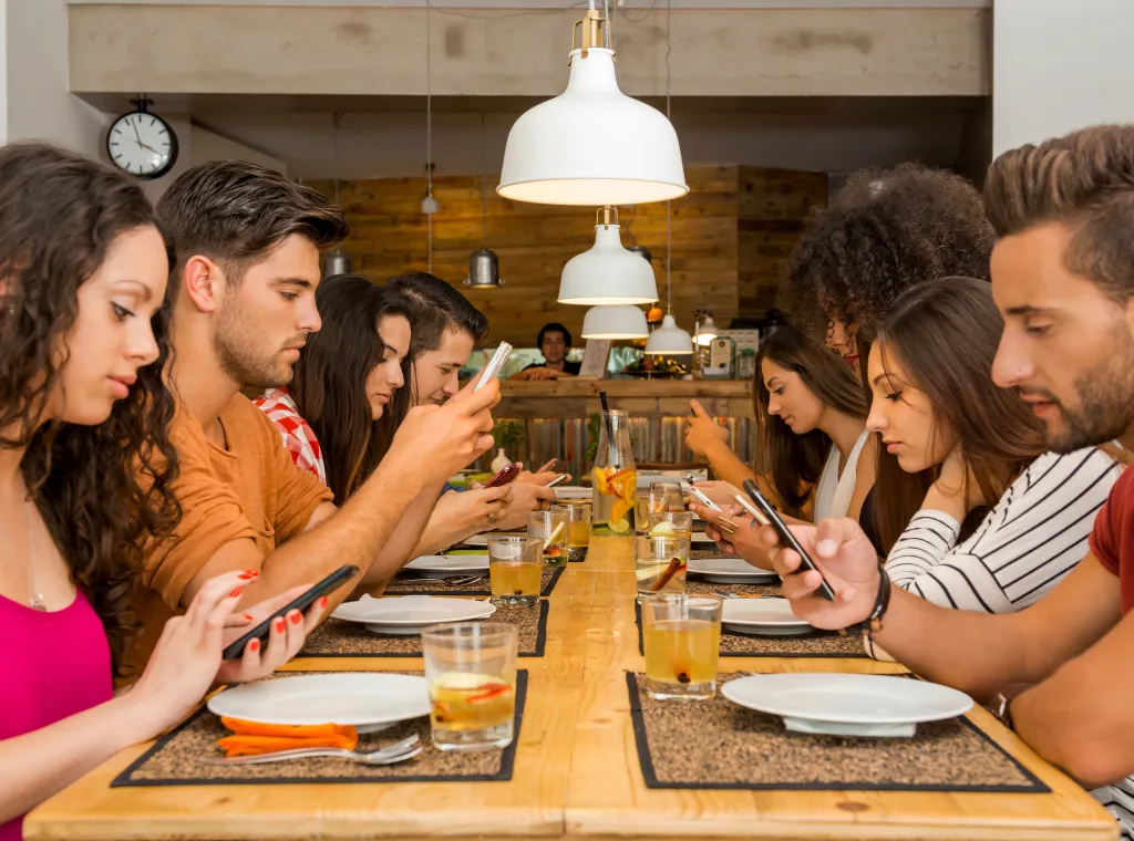 A group of young adults sit around a dining table, each looking at their phone.
