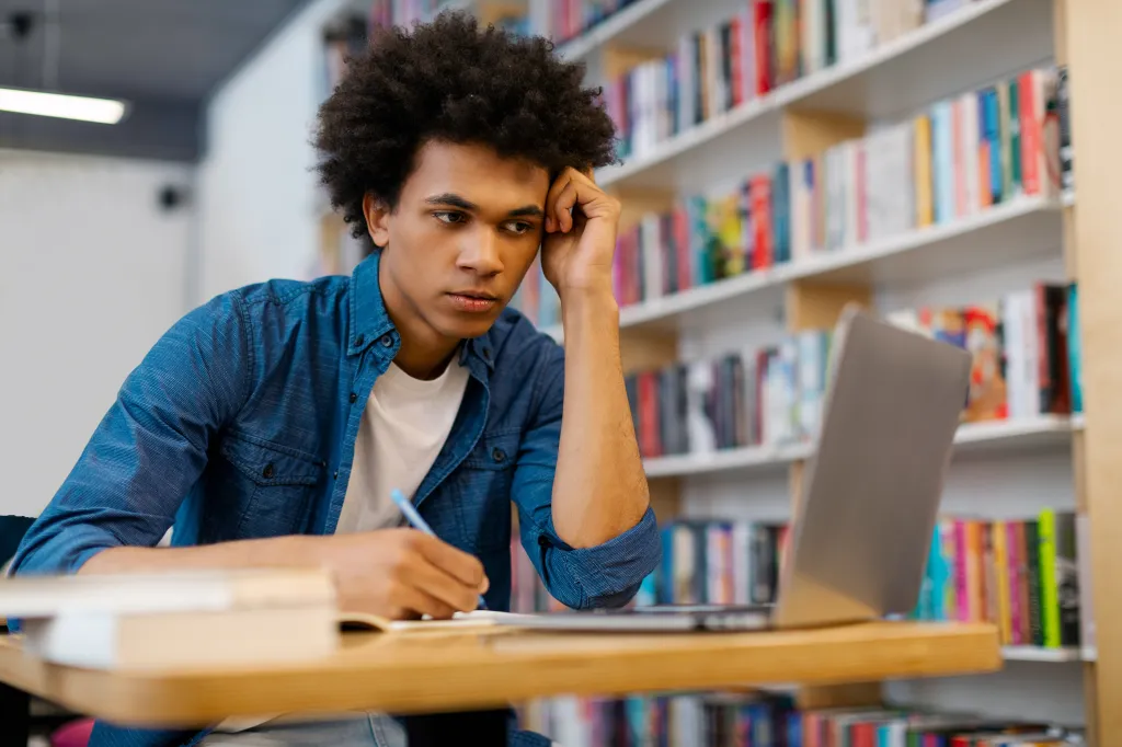 A tired male student with curly hair uses a laptop and writes notes in a library.