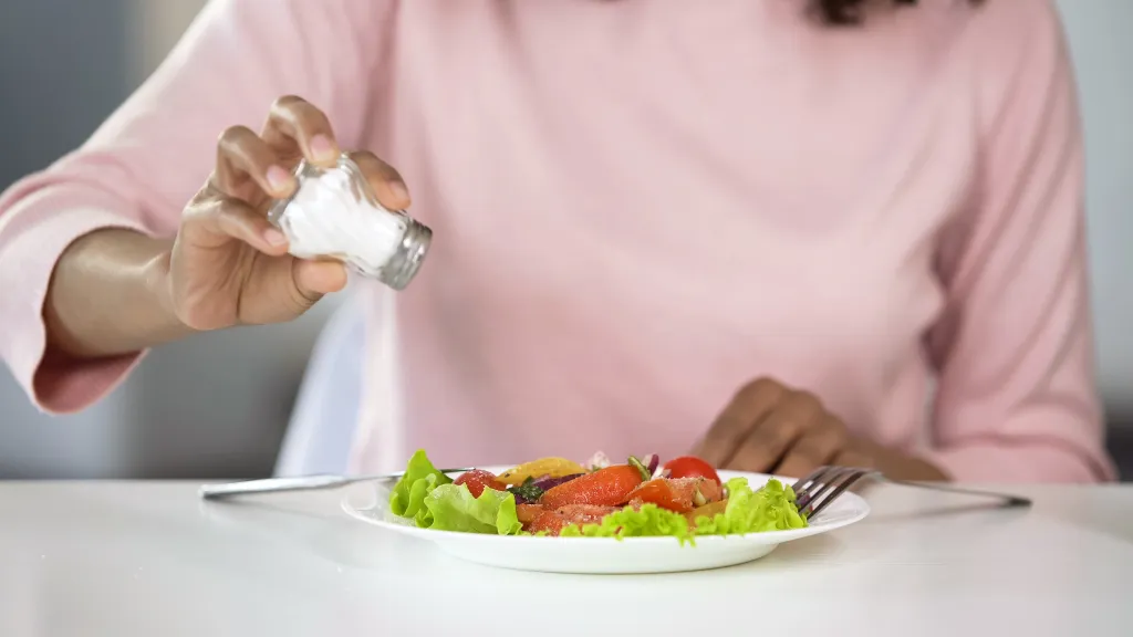 A woman sprinkles salt from a shaker onto a plate of salad.
