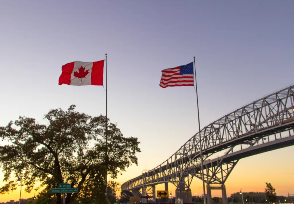 The Canadian and US flags flying side by side at sunset near the Blue Water Bridge, connecting Port Huron, Michigan and Sarnia, Ontario.