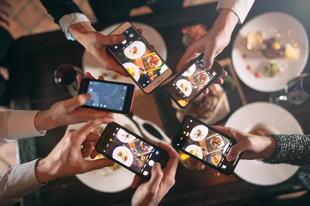 Hands holding five smartphones to photograph food on a table.