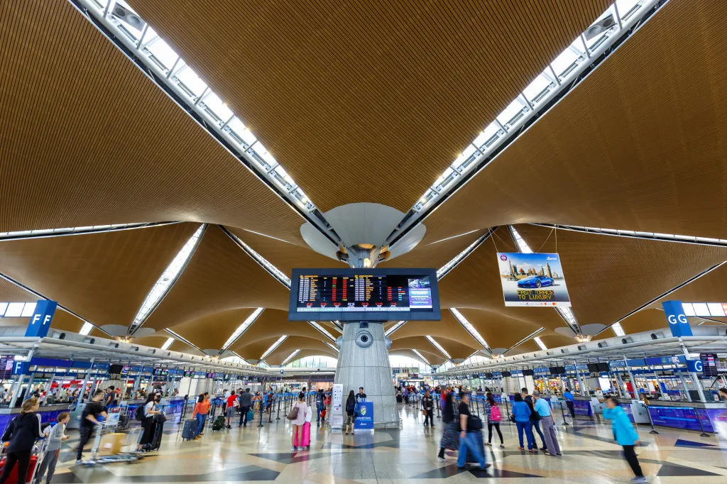 The interior of Kuala Lumpur International Airport Terminal 1, featuring its distinctive wavy, light-brown ceiling, a large departure board, and people checking in at various counters.