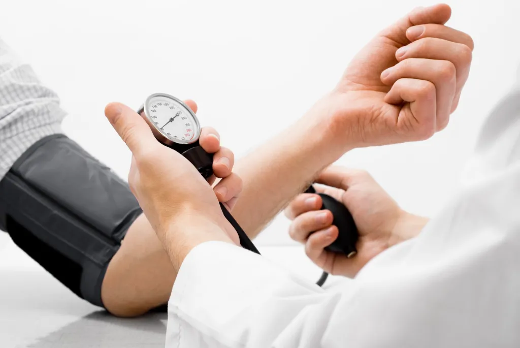 A person in a white lab coat measures the blood pressure of another person, holding the manometer and bulb in their hands.