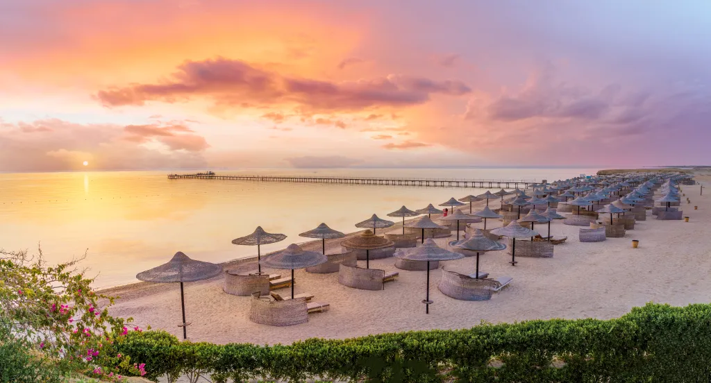 Sunrise over Fayrouz Beach Resort in Marsa Alam, Egypt, with rows of thatched umbrellas on the sand and a pier extending into the water.