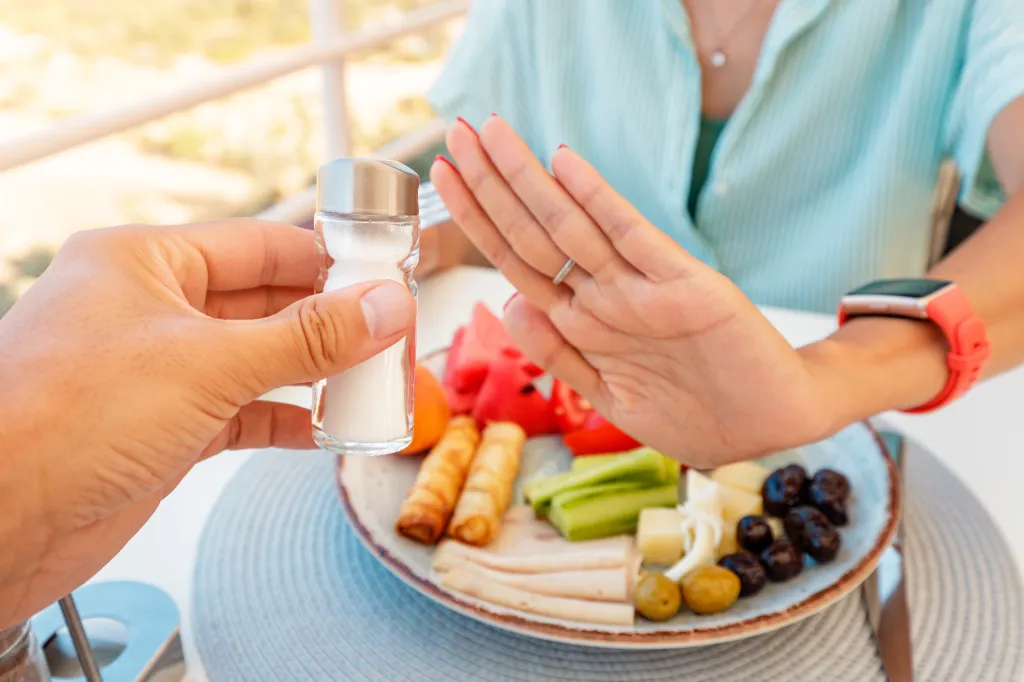 A woman refuses an offered salt shaker with her hand over a plate of food.