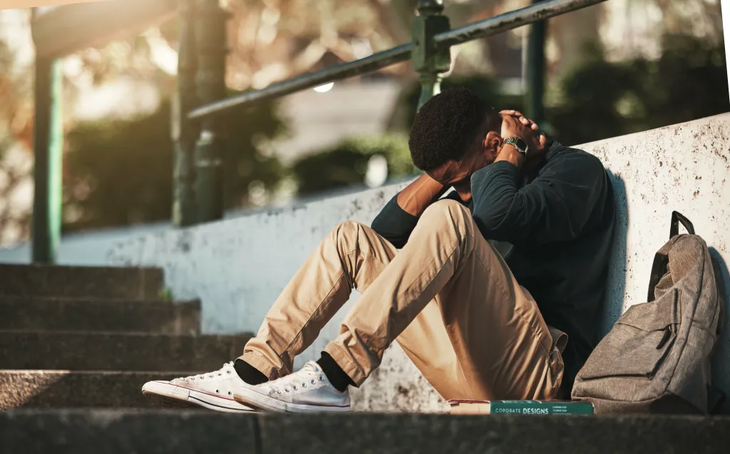 A male student sitting on concrete steps with his head in his hands, appearing stressed or upset.