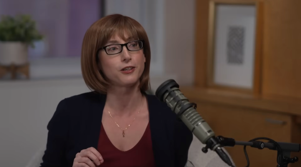 Freida McFadden with glasses and short brown hair, speaking into a microphone.
