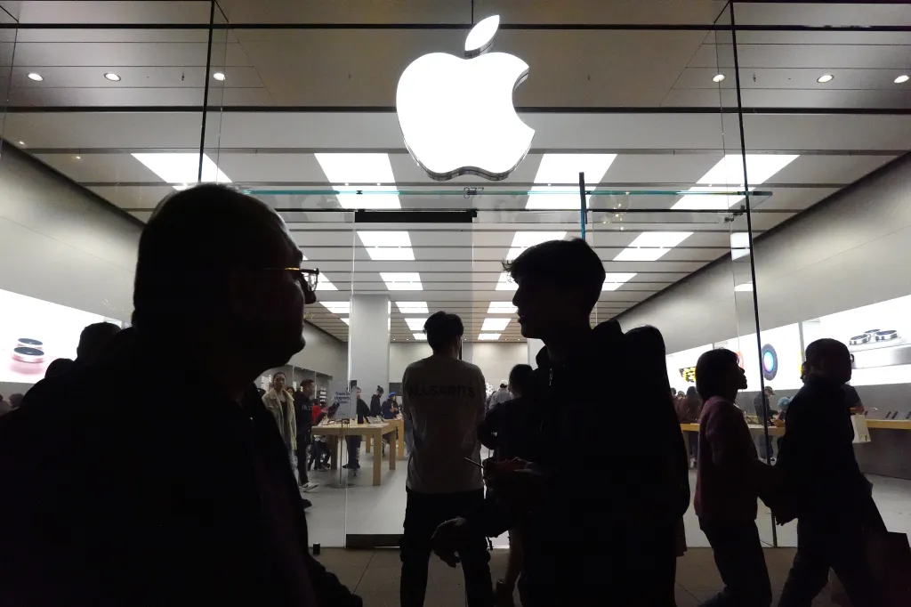 Silhouettes of people looking into an Apple Store with a large glowing Apple logo.