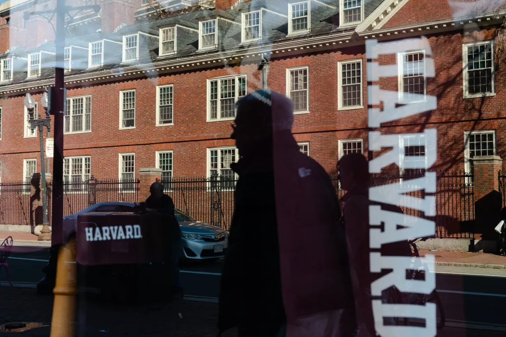 Reflection of Harvard sign and people on a storefront window with a brick building in the background.