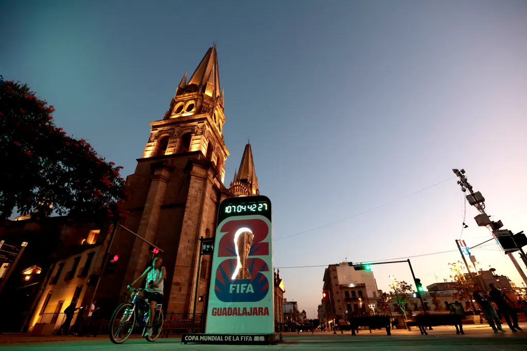 A woman on a bicycle rides past a large illuminated sign for FIFA Guadalajara and a cathedral at dusk.