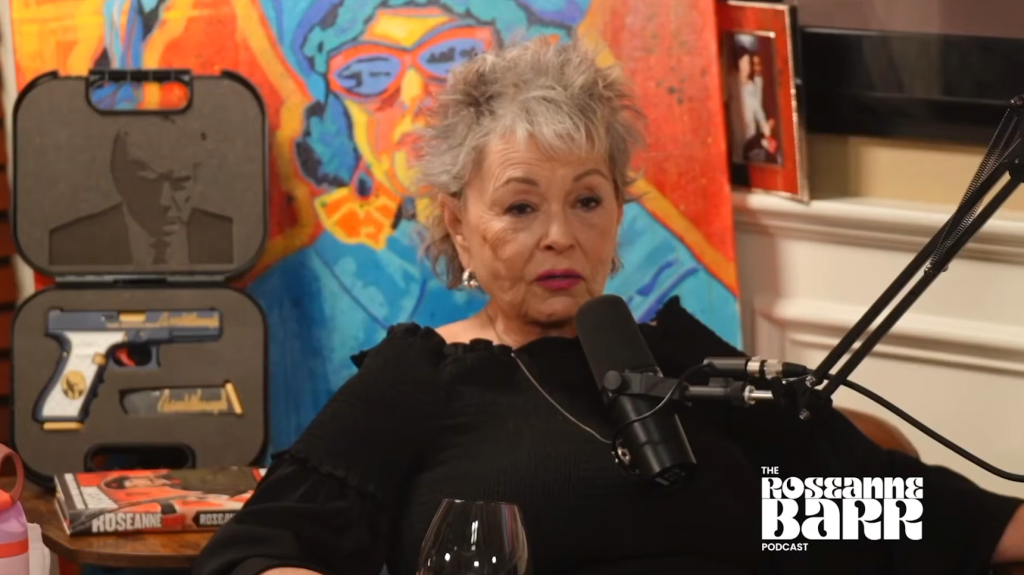 Roseanne Barr sits at a microphone with a custom gun and a Trump image behind her for her podcast.
