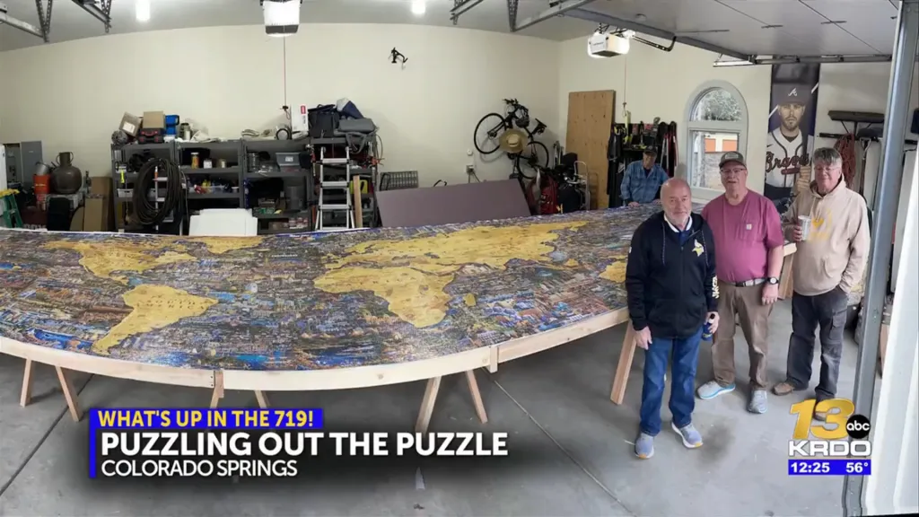 Four men stand in a garage next to a very long world map puzzle.