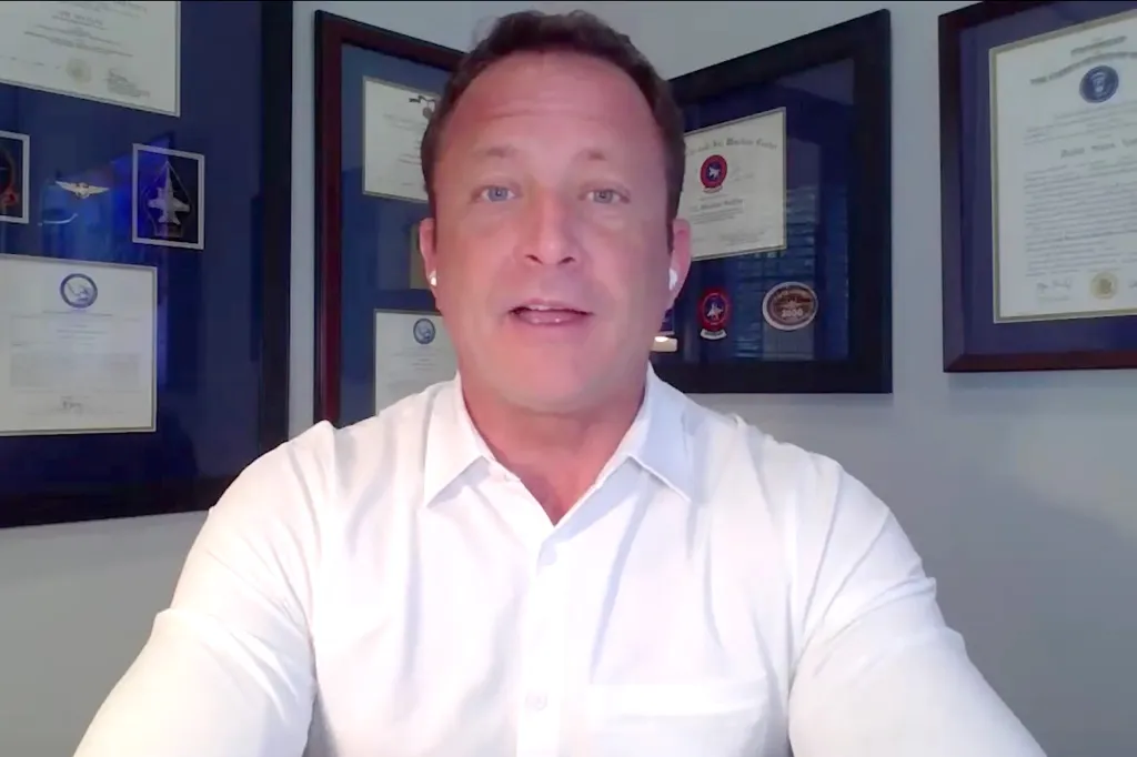 A man in a white button-up shirt speaking to the camera, with framed documents and certificates on the wall behind him.