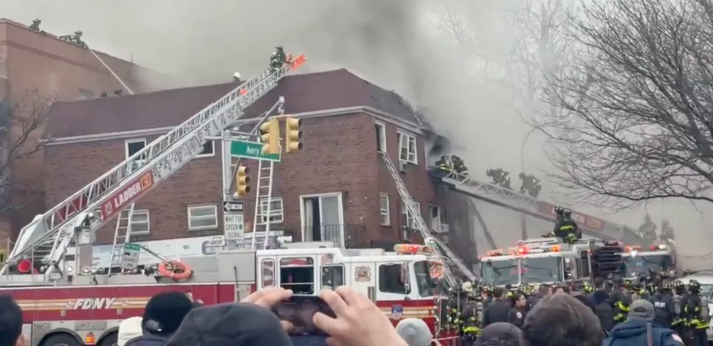 Firefighters work to extinguish a large fire at a brick apartment building, using multiple ladder trucks and hoses.