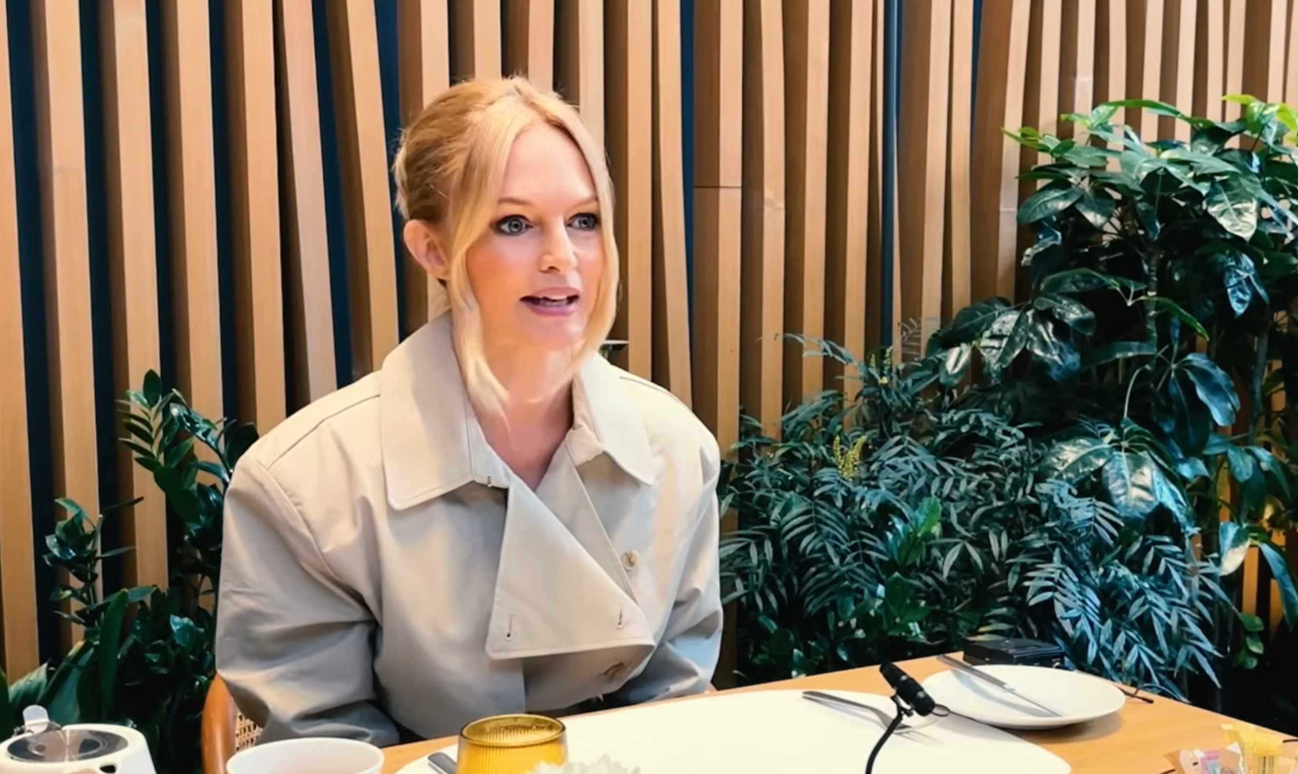 A woman in a beige coat speaking at a table with plants in the background.
