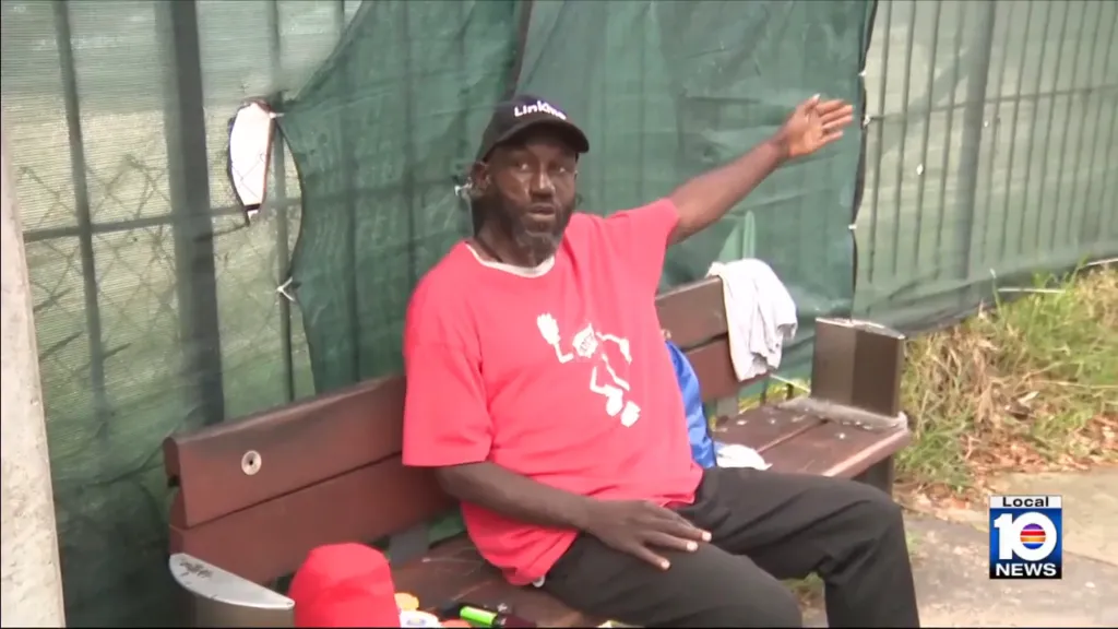 A man in a red shirt and black baseball cap sits on a bench, gesturing with his right arm.