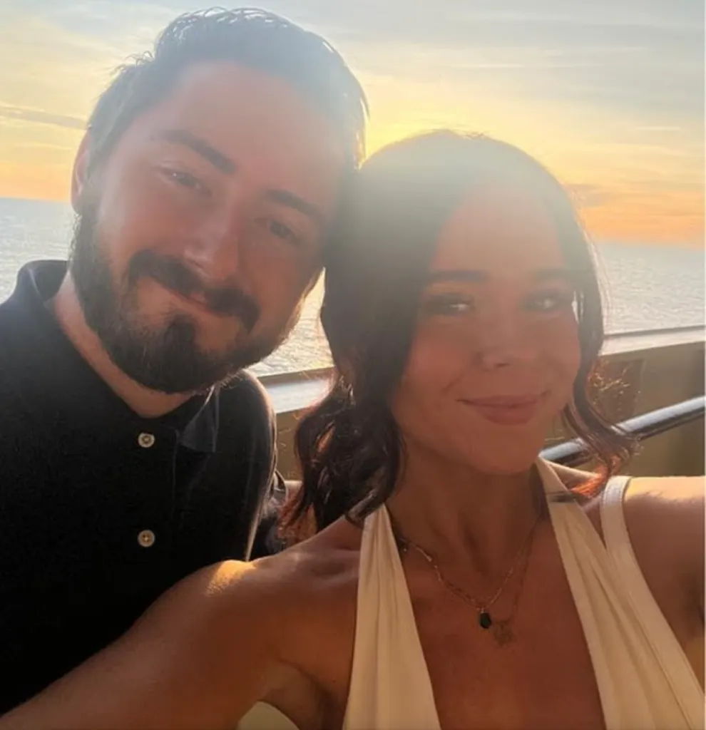A smiling man with a beard and a smiling woman with dark hair stand close together, with a sunset over the ocean in the background.