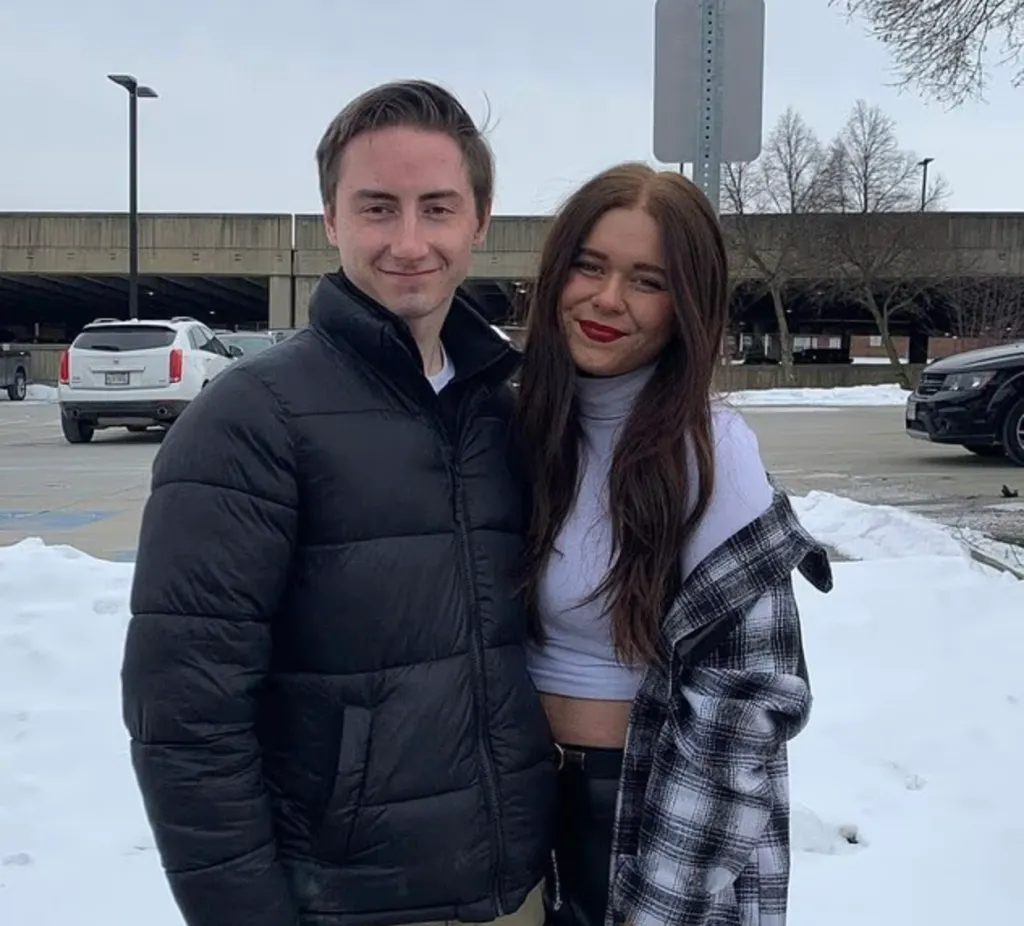A man in a black puffer jacket and a woman in a white turtleneck and plaid shirt standing in a snowy parking lot.