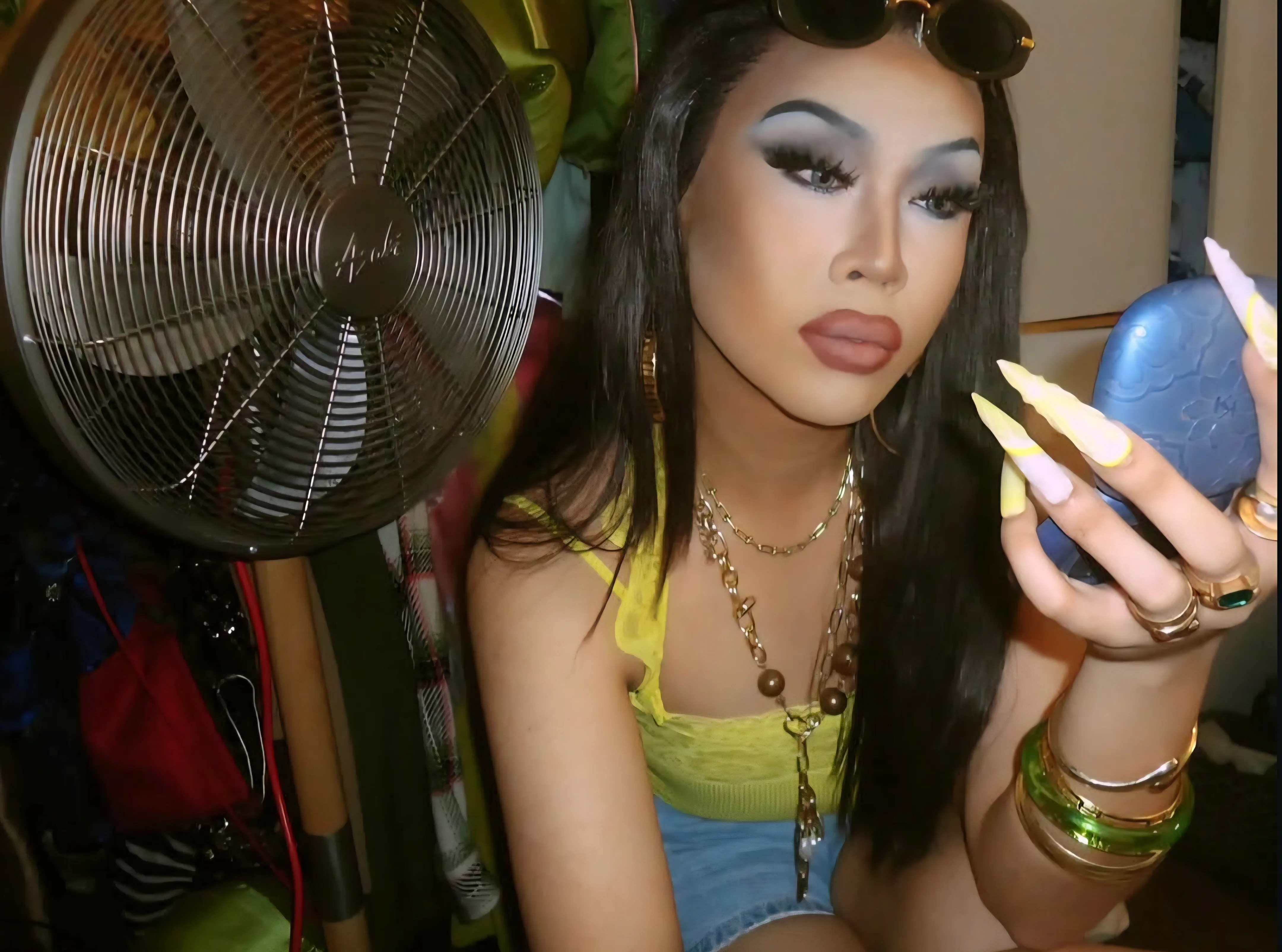 A person in heavy makeup and long nails looking into a handheld mirror, next to a fan.