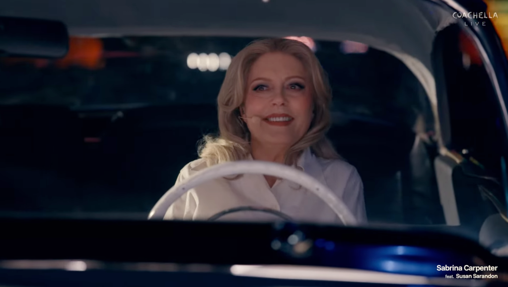 Susan Sarandon smiling in a white shirt behind the wheel of a car.