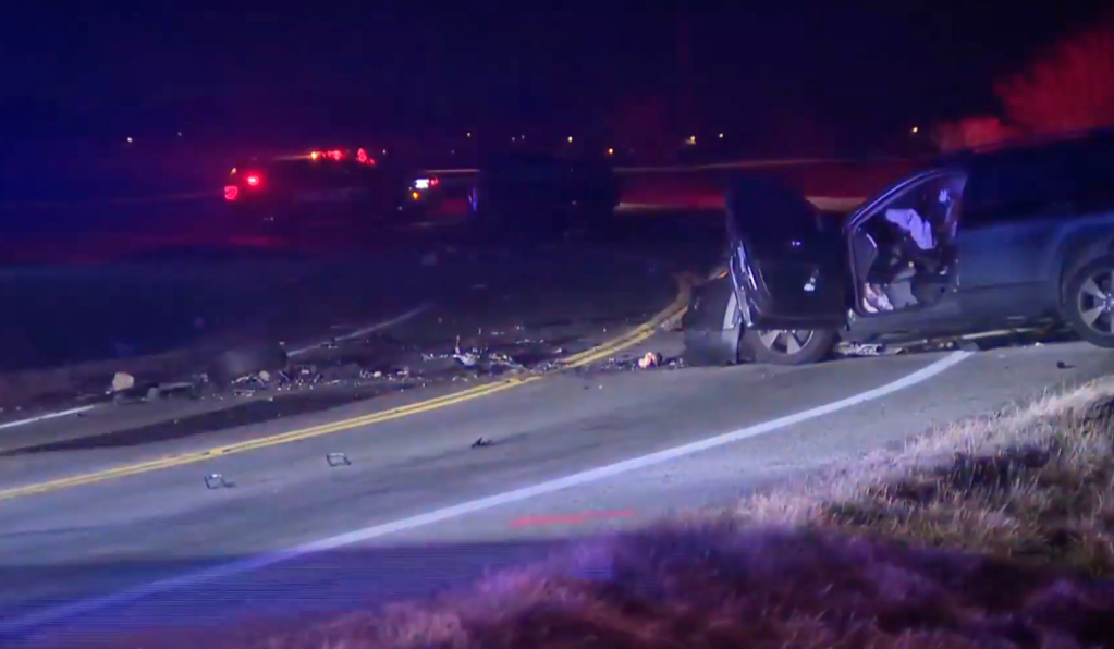 A dark blue car with doors open and debris scattered on a road at night, with flashing police lights in the background.
