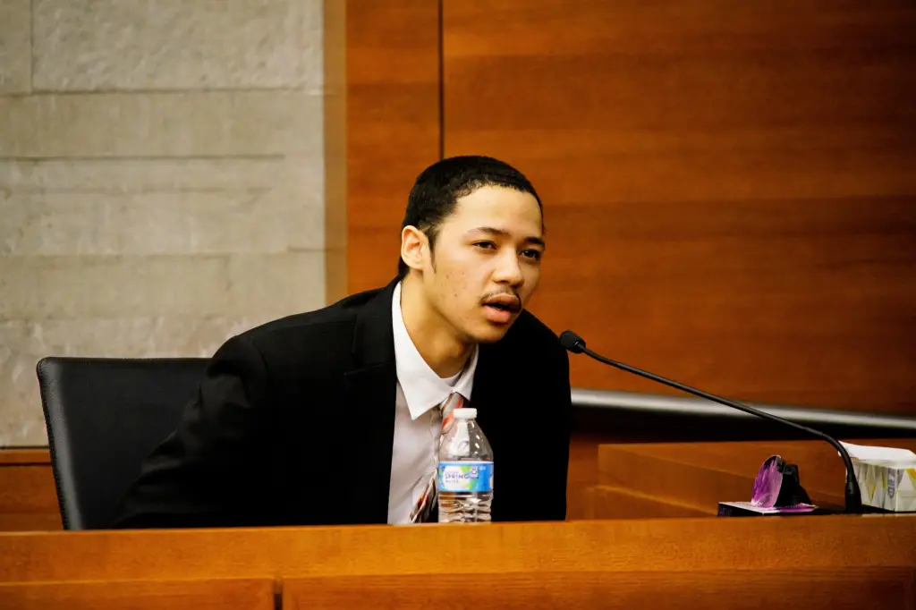 A young man in a suit speaks into a microphone at a witness stand.