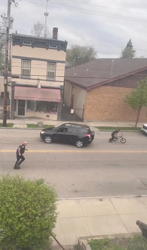 A police officer running on the street next to a black SUV and a person on a bicycle in front of a shop.