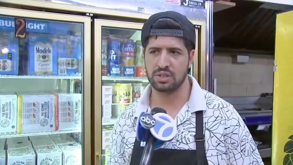 Abdul Saleh, wearing a patterned shirt and backwards cap, being interviewed by an ABC reporter in front of a refrigerator stocked with beverages.