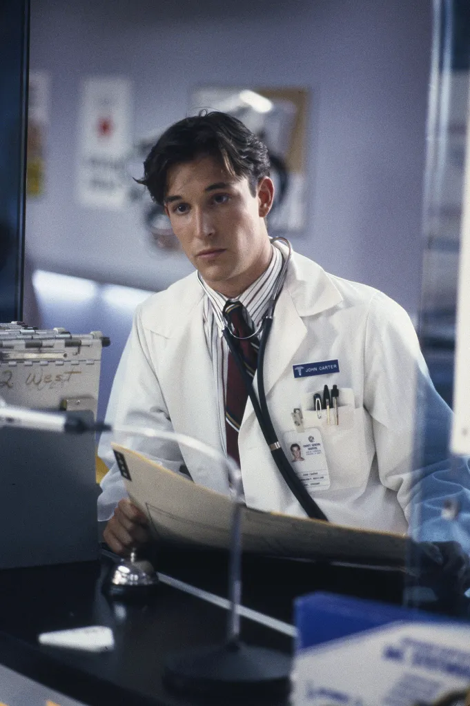 Actor Noah Wyle as Doctor John Carter in a lab coat, striped shirt, and tie, wearing a stethoscope, holding a file, and looking forward.