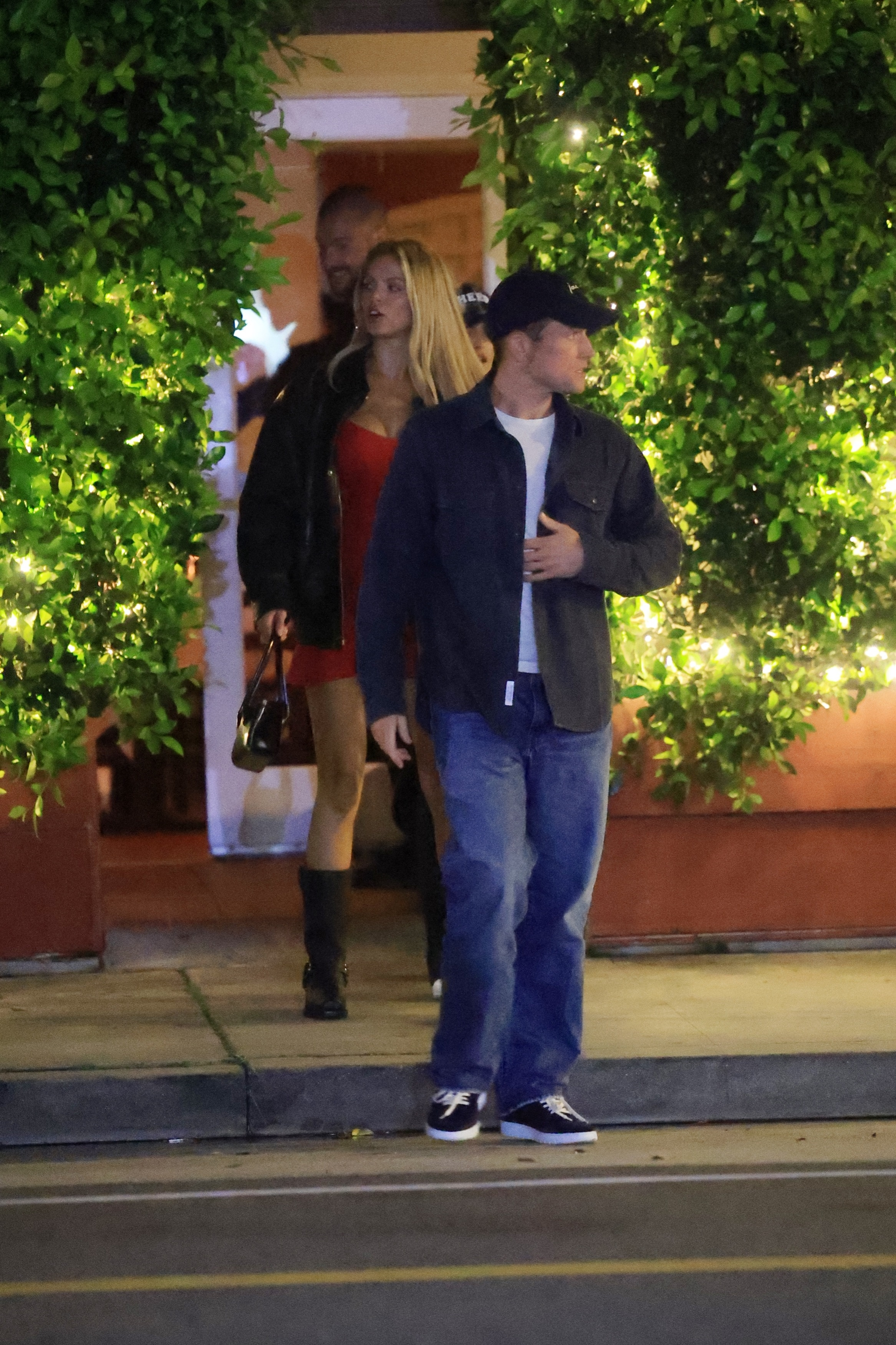 Taron Egerton in a blue shirt and jeans, and Brooks Nader in a red dress and black jacket, exiting Giorgio Baldi.